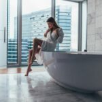 Pretty slim woman wearing bathrobe sitting on edge of bathtub filling up with water.