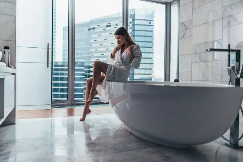 Pretty slim woman wearing bathrobe sitting on edge of bathtub filling up with water.
